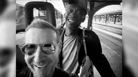 Massive Attack Massive Attack's two band members smiling in a black and white selfie at a train station. One is wearing sunglasses and the other a cap, while holding their luggage.