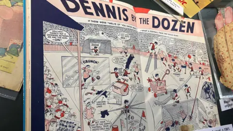 A Dennis the Menace annual from 1961 - text at the top of the double page spread in the annual reads "Dennis by the Dozen". Below it is a Dennis the Menace cartoon of a football match.