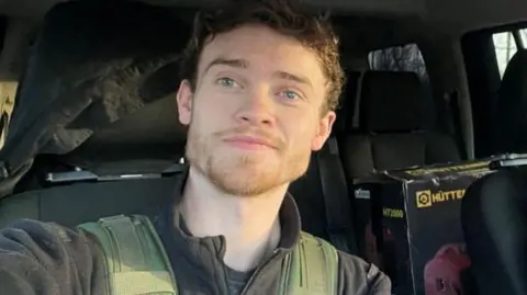Chris Parry is looking towards the camera and has his right arm stretched out as if he is taking a selfie. He is wearing a dark green fleece top with a green khaki bullet proof vest over it and is sitting in the front seat of a vehicle. There is a box of equipment on the back left hand seat.
