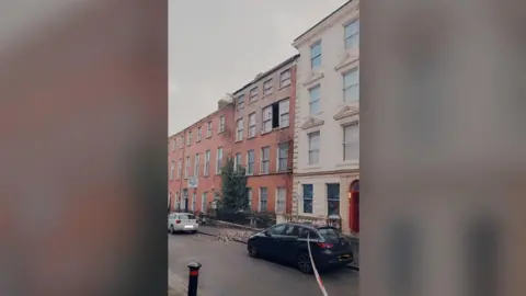 PSNI A wider view of terraced buildings on Pump Street in Derry. There is a gap between two second-storey windows at the front of a red-bricked building in the middle of the photo. There is a pile of red bricks on the pavement below. 