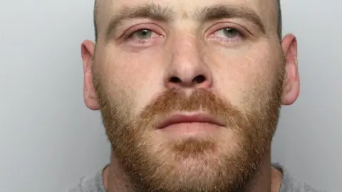 West Yorkshire Police A custody image of Mark Ross. He has a short red beard and is looking away from the camera. 