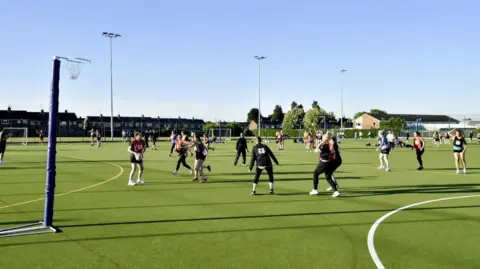 City of Lincoln Council People playing netball on a sports pitch on a sunny day. There are floodlights and a football pitch in the background.