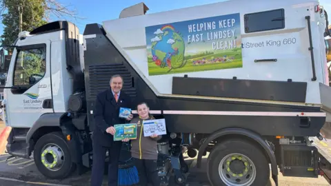 Martin Foster presenting a certificate to one of the winners. They are standing in front of one of the sweepers. The winning artwork is printed on the side with the wording "Helping keep East Lindsey clean".