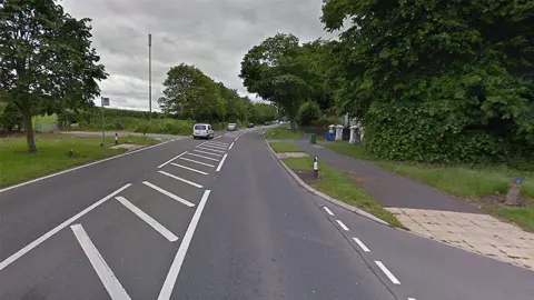 The A56 Chester Road in Walton, Warrington, Cheshire, showing vehicles travelling on the road past a bus stop with trees lining the road 