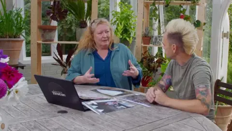 Charlie Dimmock and Lee Burkhill sitting in a conservatory at a wooden table with a laptop and a pile of photos. They are deep in conversation.