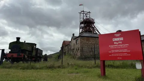A picture of the Big Pit Museum with the red welcome sign to the right and a black steam train on the left. The day is cloudy. 
