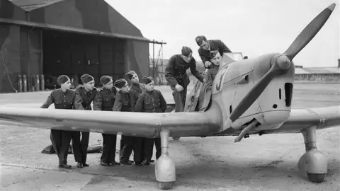 Culture Trust Luton A black and white image showing the same Miles Magister plane with a cluster of 8 cadets standing by the left wing and around the cockpit. A pilot sits in the cockpit. The plane has a number 3 on the side. In the background are large hangers which are painted in camouflage.