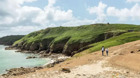 A man and a woman walking along a dirt coastal path. There are green cliffs above the sea.