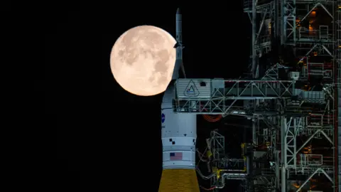 NASA A large and very bright full Moon appears directly behind the top of Nasa's Space Launch System rocket at night. The orange and white rocket is sitting next to a steel support structure called a mobile launcher.
