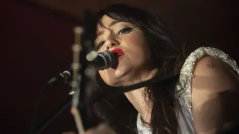 Singer KT Tunstall with long dark hair and a silver top sings into a microphone