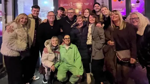 Jade Franks Jade in a green tracksuit is crouching in front of a large group of people. They are all smiling at the camera with their arms around each other. 