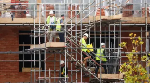 Four men climb down scaffolding stairs on the side of an under-construction brick building. They are wearing yellow high-vis jackets and hard hats.

