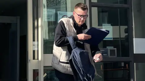 PA Media Steven Carruthers leaving court. He has dark hair and is wearing glasses, a dark top and a cream padded gillet. He is carrying a blue rucksack and is holding a blue folder.