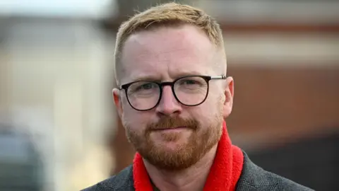 Lloyd Russell-Moyle. A man with short ginger hair and a beard looking at the camera. He is wearing black round glasses and a red scarf