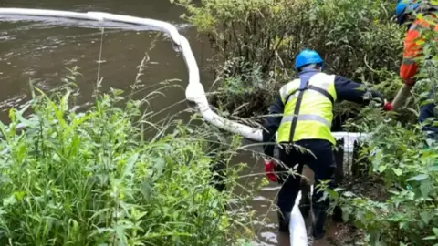 A helmeted worker stands in a river next to a floating bund which is being used to contain oil