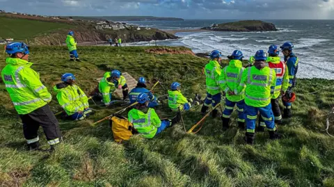 Kingsbridge Coastguard Rescue Team A large group of HM Coastguard search and rescue team members in high-visibility jackets gather on a windswept cliff above a rough sea, appearing to carry out a coordinated training exercise or rescue operation with ropes and equipment.