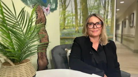 Rachel Beckett sitting next to a table. She has blonde hair and is wearing black-rimmed glasses, a black top and black suit jacket. She is looking into the camera. A plant is in the foreground of the picture on the left-hand side.