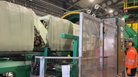 Several large white containers are on a conveyor belt. The end of one of them is open and a huge pile of rubbish or recycling can be seen inside. There is a very tall barrier besides the conveyor belt. A man wearing a blue cap, yellow ear defenders and an orange high vis jacket with reflective silver stripes on it.