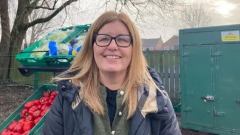 A woman with shoulder-length dark blonde hair and glasses, stands in front of food pantry stalls in a primary school playground. She wears a black puffer jacket, and a green cardigan.