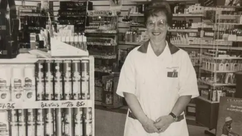 An old, black and white picture of Sadie in her work uniform in the middle of the chemist.