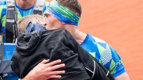 PA Media A man in a blue and green patterned top and headband carries a man wearing a black top in his arms and kisses the side of his face. Behind them is another runner in a green and blue top.