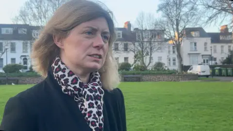 BBC Deputy Feltham is wearing a black coat and has a white scarf with ref flowers. 