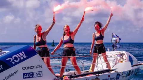 The three women are standing in their boat holding up red flares as they celebrate finishing their row.