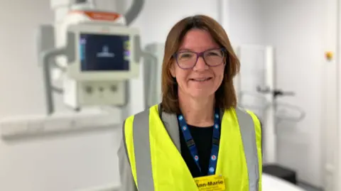 A woman with shoulder-length brown hair, and glasses. She is wearing a high-vis vest over her clothing, and has a yellow name badge. There is medical equipment behind her