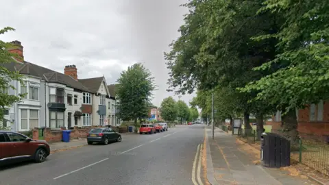 Hainton Avenue in Grimsby. It is a wide road with a pavement and houses on either side. There are a number of park cars in the image, as well as trees and a black bin.