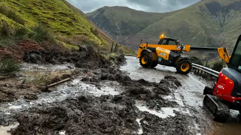 Soil from a landslip on the road, with the verge to one side and clean-up vehicles on the other