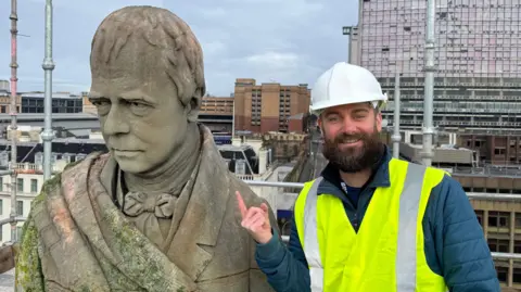 A large statue of Sir Walter Scott. Beside it, a bearded man in a white helmet and yellow hi-vis tabard is smiling and pointing at the statue. Buildings can be seen in the background, as can scaffolding.