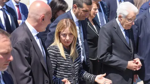 Getty Images Prime Minister Giorgia Meloni and President Sergio Mattarella pictured walking amongst a group of men and women in suits. Meloni, with blonde hair past her shoulders, is wearing a smart striped jacket and is gesticulating with her hands while speaking to the man next to her. Mattarella, with white hair and glasses, has his hands clasped together and is looking straight ahead.