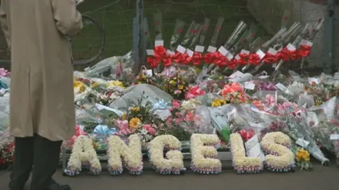 Getty Images A pile of flowers, some of them spell out the word ANGELS in capital letters. They are all propped against a metal grid fence. A man in a brown raincoat is standing with his back to us, looking at them.