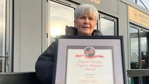 Tracy Howard has short grey hair and is standing in front of a building which has a sign saying "Marine in the community". She is holding a framed "longest serving league manager award" certificate presented to Roly Howard in 1997.