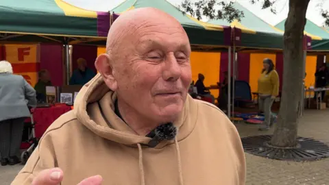 A man in a beige hoodie is looking just past the camera a smiling. Behind him are green, yellow and purple marquees.