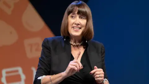 Jane Dodds delivers a speech at the Lib Dem party conference. She has a dark brown bob and fringe and is wearing a black dress with a black blazer over the top. 