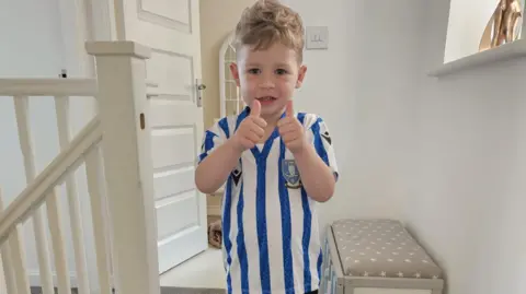 A smiley little boy wearing a blue and white striped football shirt holds two thumbs up to the camera.