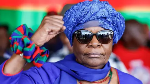 AFP Netumbo Nandi-Ndaitwah wearing black sunglasses and a blue patterned scarf raises his fist in victory.