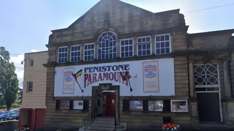 Streetview image of the cinema. It is a small but grand-looking stone-built building. A large horizontal banner covers the middle third of the facade, and says "penistone paramount" in a stylised font. There are pride flags either side of the door.