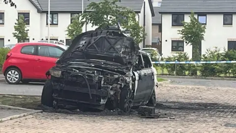 A car is black with soot. It has been burnt out. It's bonnet is flipped open and everything is black or melted. there is a red car in the background and white  houses.
