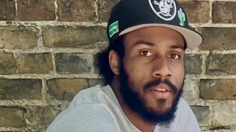Close-up photograph of Auguste-Marie Bile-aka Amoa with a full dark beard and moustache looking directly into the camera. He is wearing a light grey T-shirt and a black Las Vegas Raiders baseball cap featuring the team logo on the front and a green patch on the side. He is posing against an outdoor background of weathered brown and beige brickwork with light mortar.