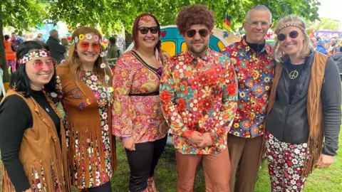 BBC/Oli Constable A mixed gender group of people, dressed in 1960s hippy-style fancy dress stand together smiling.