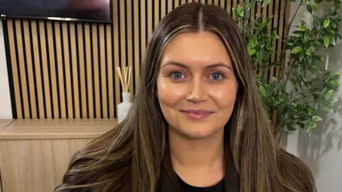 Codie Foster is smiling and has long, brunette hair with blonde highlights. She is wearing a black top and black blazer. Behind her is some light-coloured wooden office furniture and a green plant. 