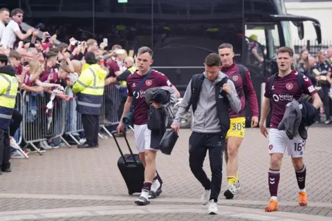 PA Media Hearts players arrive at the stadium carrying bags as fans gather behind barriers to greet the team before the match.