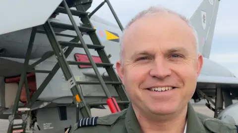 A man wears a dark green RAF pilot uniform and smiles into the camera. He is standing in front of a Typhoon fighter jet. The man has short grey hair and dark blue eyes.