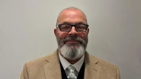 A man with a silver and grey beard wearing black-rimmed glasses and a light brown jacket and white shirt and light silver/gray tie. 