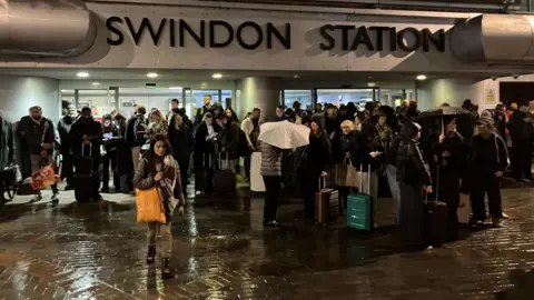 Dozens of passengers are gathered outside Swindon Station at night. Rain is falling and the large Swindon Station sign is visible above the people