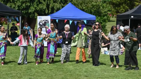 Eid celebrations taking place at Northampton’s Becket’s Park. People are holding hand and standing in a line while standing on the grass. 



 