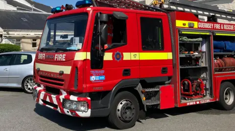 BBC A red Guernsey fire and rescue fire engine outside in a built up area with blue skies. The side panels where the hoses are kept is open. There is a silver car next to the truck. 