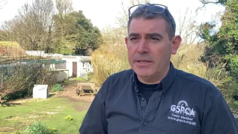 GSPCA manager Steve Byrne stands in a garden in front of a set of trees, a couple of park benches and a white single-storey building which has a brown door. He is wearing a grey shirt and has his glasses placed on top of his head.